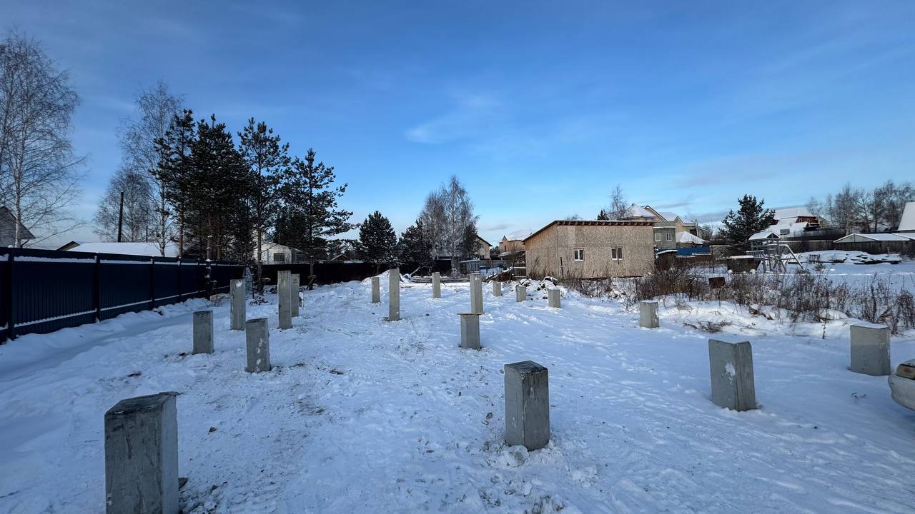 Снежный участок в посёлке Красный с рядом забитых железобетонных свай на фоне заборов и деревьев.