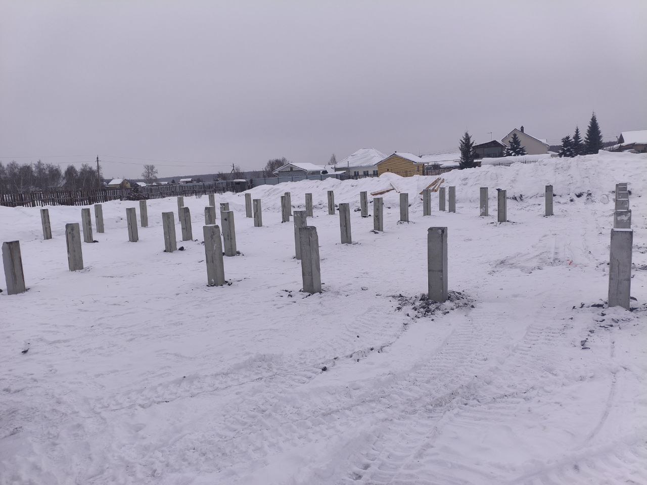 Забитые ЖБ сваи на подготовленной площадке зимой, видны следы техники и соседние постройки в селе Покровском