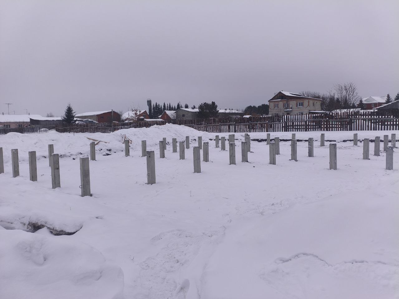 Свайное поле из железобетонных свай С40.20-у на заснеженном участке в селе Покровском, вид на частные дома и забор по периметру
